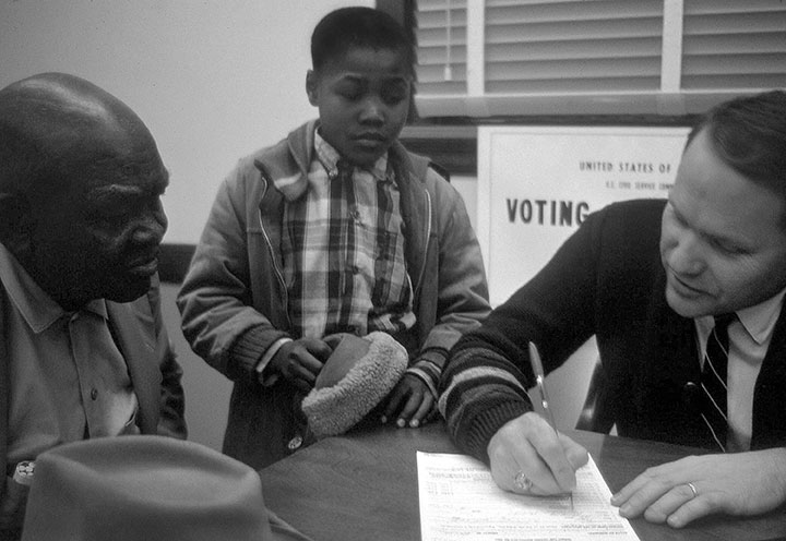 A federal agent enrolls an elderly black man to vote for the first time in Mississippi as a young boy looks on, Tylertown, MS 1965-­‐6